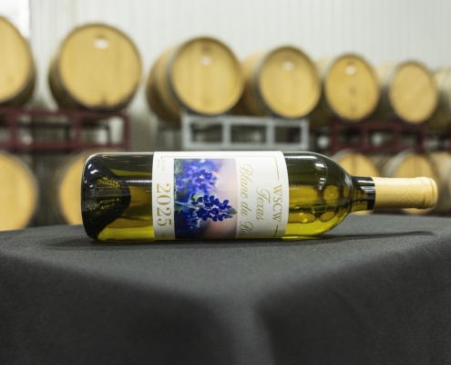 A bottle of WSCW 2025 white wine lays centered on a table, set inside a winery with large wine barrels in the background.