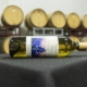 A bottle of WSCW 2025 white wine lays centered on a table, set inside a winery with large wine barrels in the background.