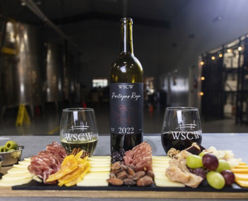 A bottle of WSCW 2022 red wine stands centered between two branded wine glasses, one white and one red, behind a charcuterie board with sliced cheeses, cured meats, nuts, dried fruit, olives, and grapes, set inside a winery with large stainless steel tanks in the background.