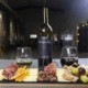 A bottle of WSCW 2022 red wine stands centered between two branded wine glasses, one white and one red, behind a charcuterie board with sliced cheeses, cured meats, nuts, dried fruit, olives, and grapes, set inside a winery with large stainless steel tanks in the background.