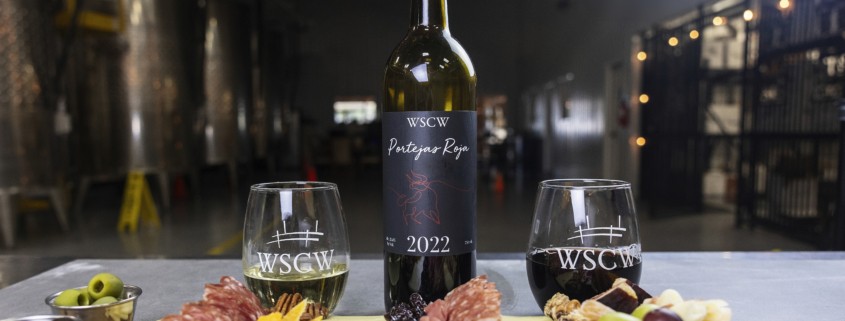 A bottle of WSCW 2022 red wine stands centered between two branded wine glasses, one white and one red, behind a charcuterie board with sliced cheeses, cured meats, nuts, dried fruit, olives, and grapes, set inside a winery with large stainless steel tanks in the background.